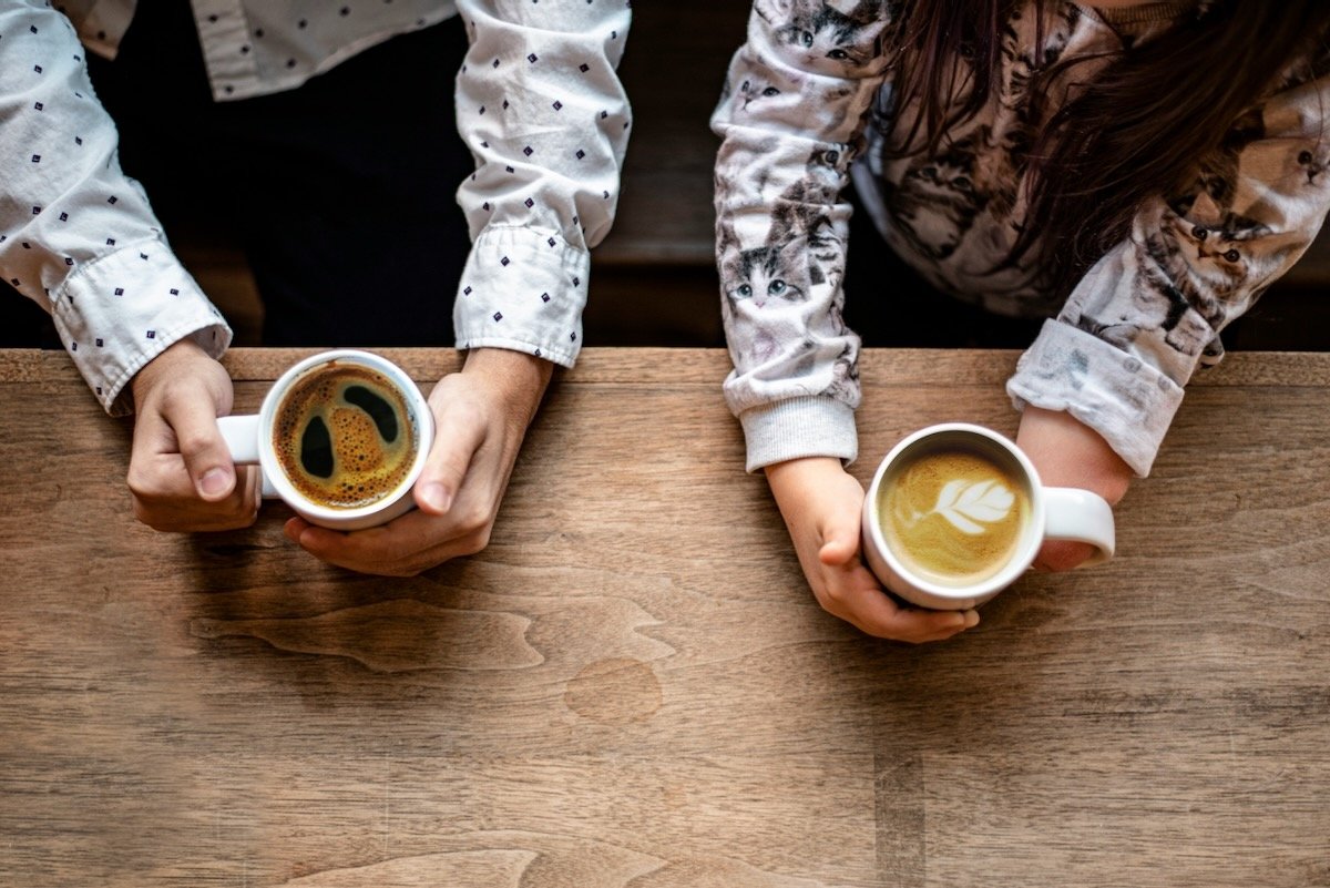 Two people holding cups of coffee and latte at a wooden table, photographed from above.