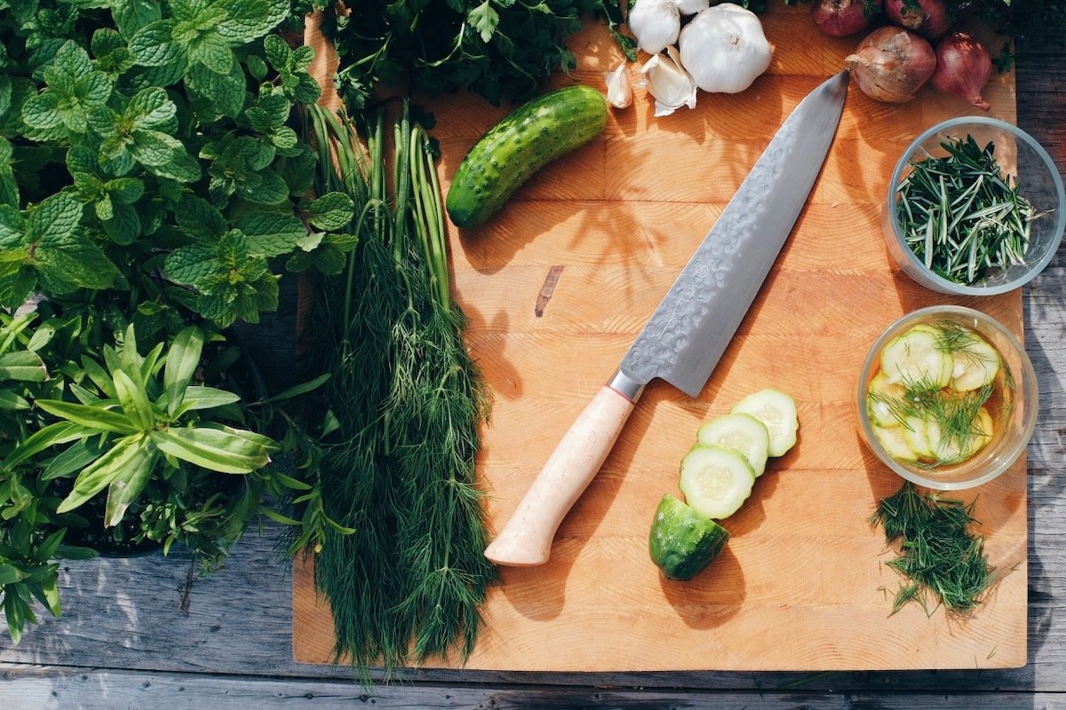 Fresh herbs, cucumber, garlic, and a kitchen knife arranged on a wooden cutting board.
